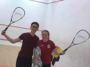 Ellie with a friend, both holding up their racquets and smiling, in the Christ's College squash court